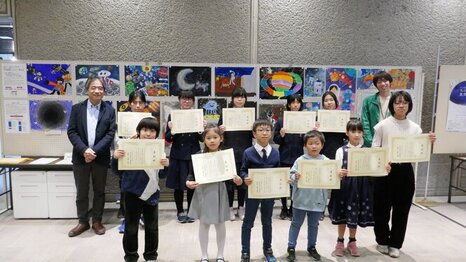 令和6年「宇宙の日」記念行事 全国小・中学生作文絵画コンテスト受賞者と館長との記念写真。賞状を持った受賞者が掲示してある作品の前で並んでいる。